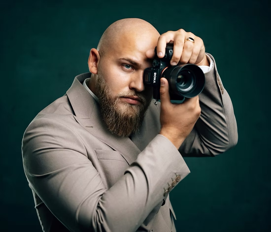 Fotograf und Filmemacher Alex Kardos im sandfarbenen Anzug blickt durch seine Kamera
